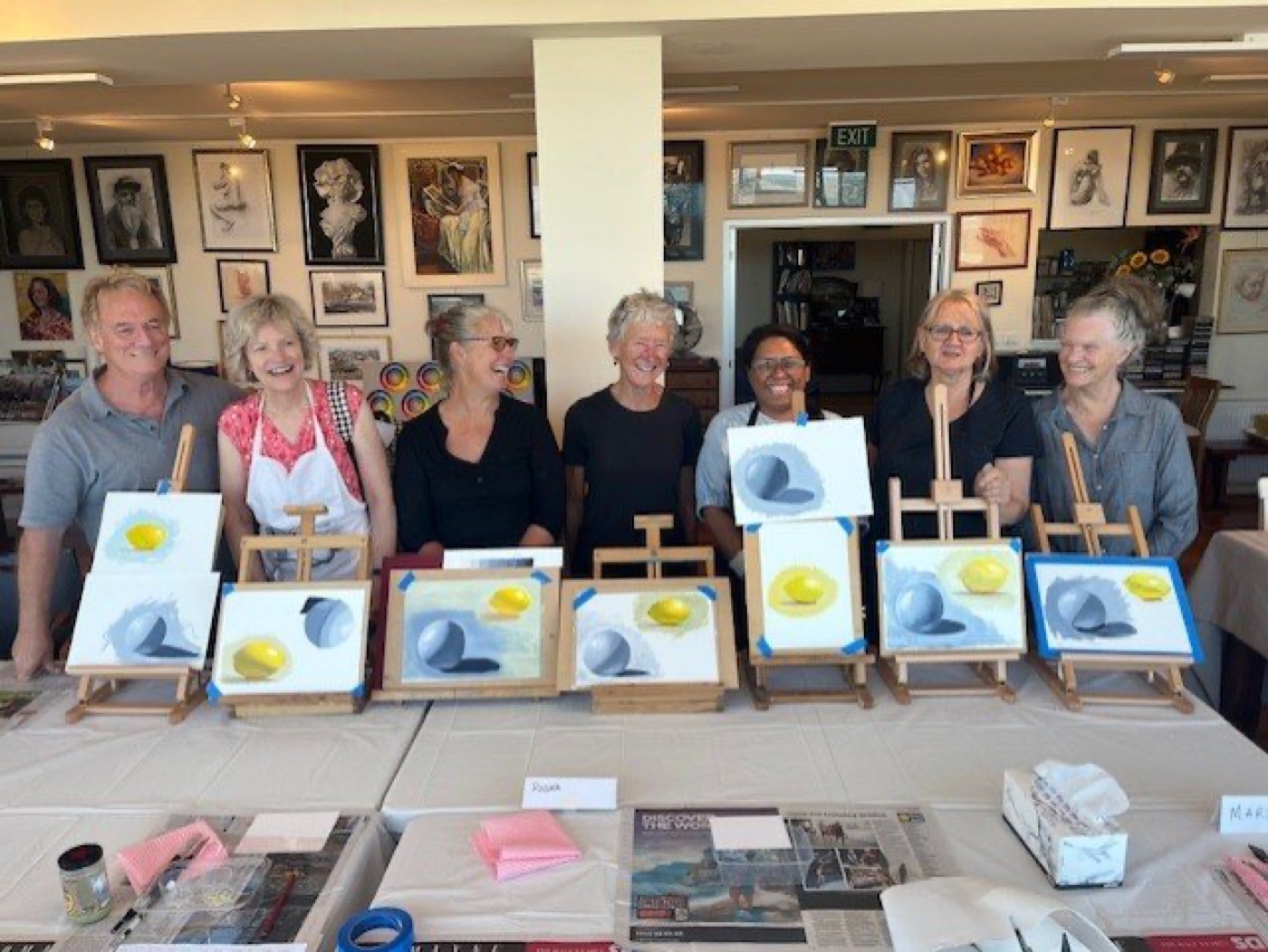 David Payne and seven students standing behind their easels at the end of an oil painting workshop, gallery wall of framed art behind them